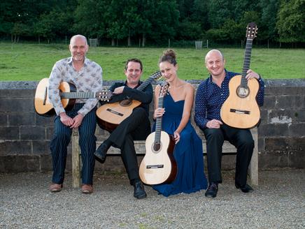Three men and one woman sit on a bench in front of a field. They are all holding guitars.