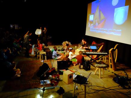 A group of children are sat in front of a movie screen with instruments like shakers, cymbals, recorders, guiros, a gong and xylophone..