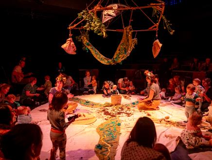 A group of children and their parents are sat in a circle alongside a guitarist. Two other adults sat inside the circle, engaged with the children.