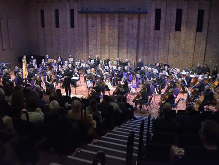 An orchestra is seated on the Turner Sims stage with an audience watching.