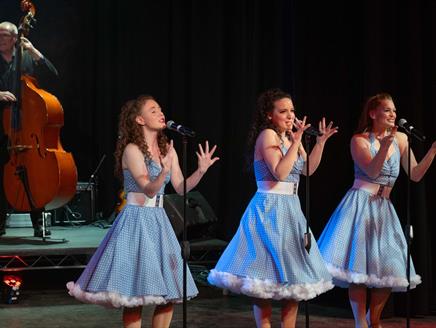 Three women dressed in blue 1950s style dresses singing with a cello player in the background.