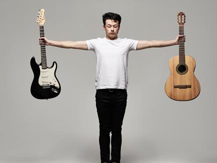 Sean Shibe stands holding two guitars, an electric guitar to the left and an acoustic guitar to the right