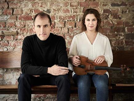 Chloë Hanslip and Danny Driver look towards the camera with a brick wall behind them.