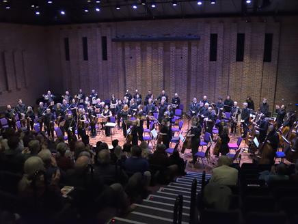 The image has a full orchestra standing on Turner Sims' stage. The orchestral players and conductor are looking towards the audience.