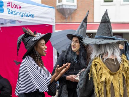 Three ladies dressed in whitches hats are taking to each other. Text on a pink gazebo in the background reads "Love Basingstoke".