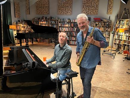 One man sits at a piano while another man stands next to him, holding a mezzo-soprano saxophone and smiling.