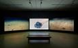 Image of Emma Critchley's film work Soundings, showing the deep sea and associated matter over three separate screens. The room is dark and there is a