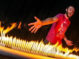 A man with a pink T-shirt on is standing in front of a large metal tube that has lots of holes in it. There is fire coming out of the holes and the ma