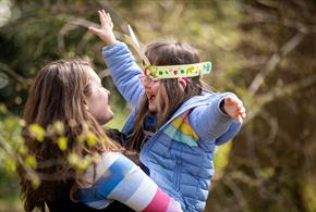 A parent lifts their child up. The child wears bunny ears and a lilac coat and has their arms up in the air.