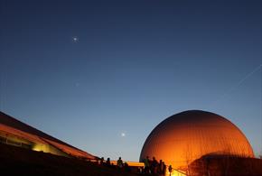 A dark sky with a few stars. The Science Centre and Planetarium are glowing orange against the night sky.