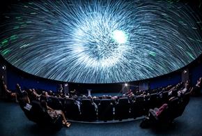 A blast of white light on a Planetarium dome. There are people sat in seats watching the screen.