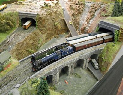view of a model railway from above. Two trains are crossing a bridge, coming out of a tunnel.