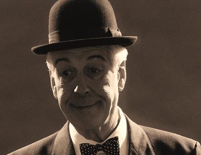 a black and white photograph of a white man, showing his head only. He wears a bowler hat, bow tie and has painted on eyebrows.