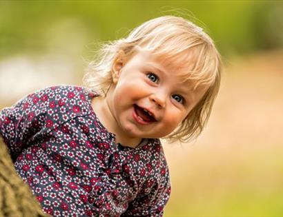 a photograph of a small white child with blonde hair smiling as they appear behind a tree trunk. background is blurred.