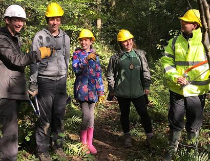 a photograph of five people stood in forest with hard hats on. man to right wears high viz jacket whilst others are young adults / teenagers.