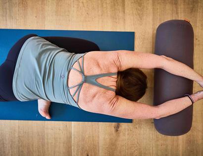 Person practicing a supported yoga stretch on a mat, arms extended overhead and resting on a bolster in a calm indoor setting.