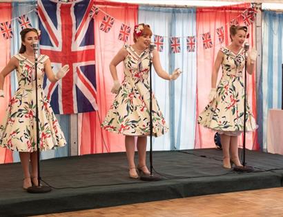 three women on stage in vintage swing dressers singing. Hair in 1940s style. Union jack flag and bunting in background.