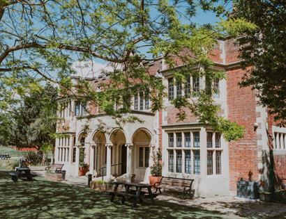 Exterior of Hastings Museum & Art Gallery, a red-brick building set in gardens with trees and picnic benches.