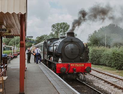 Steam locomotive WD75008 at a Kent & East Sussex Railway station platform, with smoke rising overhead.