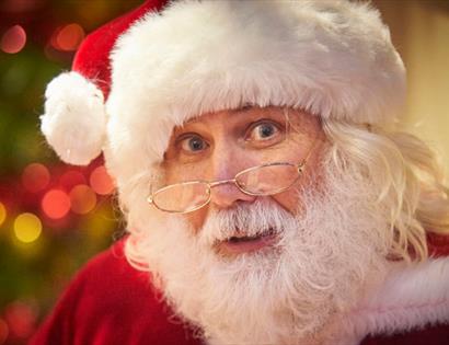 photograph of father christmas. A close up of a man witha white beard and moustache with red outfit and white-fur lined hat, spectacles reading on nos