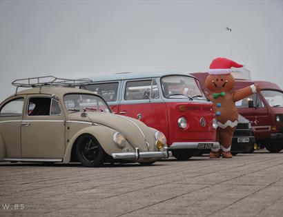 four vintage volkswagens, with beetle in the foreground and three campervans behind. A giant gingerbread character is standing next to them.