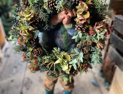 a small child holding a door wreath to the camera, their blue boots visible but their face completely hidden by the wreath.