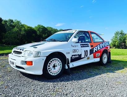 a white rally car with racing stickers over it, parked on gravel with green lawn and blue sky in background.