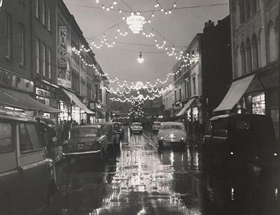 black and white photograph of street scene with 1950s cars and festoon lights reflecting in wet street