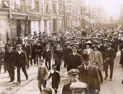 black and white photograph of crowds walking down terraced street, mostly wearing smart clothes and caps.