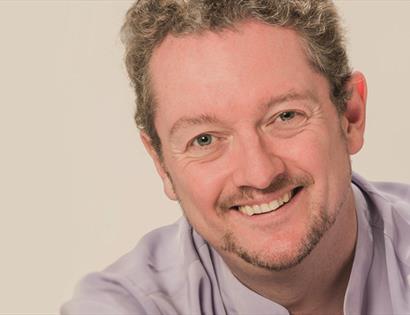 a portrait of a white man, smiling, just visible lilac shirt against a light plain background.