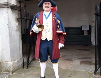 Town crier dressed in traditional dress, burgundy and blue with a tricorn hat with red feathers, holding a large bell.