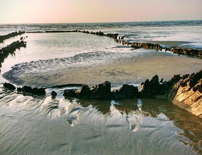 a photograph of the wreck the amsterdam. shows wet sand at very low tide with outline of boat emerging from it.