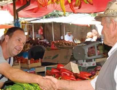 A market seller shaking hand across his veg stall with a man wearing a hat.