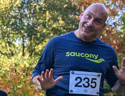 Runner wearing bib raises both hands while crossing a forest trail during a run at Bedgebury National Pinetum.