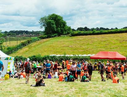 Outdoor event with people gathered under colourful tents on a grassy field with trees and hills in the background.