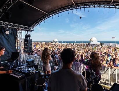 Photograph taken from within outdoor stage, look out at large crowd with the sea in the background.