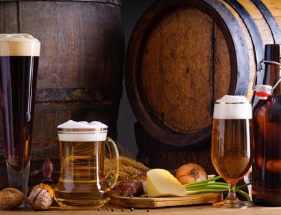 Photograph of three glasses of ales, with wooden barrel in background, surrounded by vegetable and cheese produce.