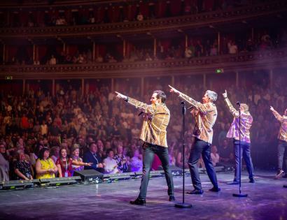 A photograph of musical theatre group Collabro performing on stage. The back of four men in gold jackets in a packed theatre. They each point to the s