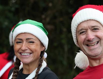 Two people in festive hats smiling at camera outdoors, promoting the Christmas Pudding Dash fun run event.