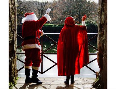 backs of father christmas and woman in red cloak waving out of castle archway.