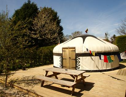 A yurt on decking with a bench in front. There is blue sky and bunting.