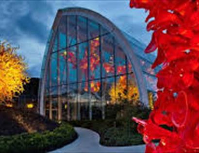 photographs shows cotemporary glass house at dusk with coloured glass sculptures in foreground, looking like trees.