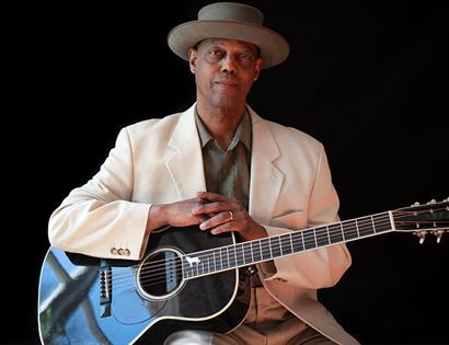 Photograph of balck man wearing a white suite jacket and brimmed hat with arms placed on his guitar. Against a black background.