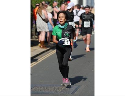 Runner in black and green Hastings Runners shirt with race bib on a sunny street during a 10K event.