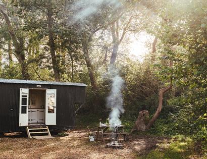Shepherd’s hut by The Original Hut Company near Bodiam, with a smoking fire pit and wooden seating in a woodland clearing.