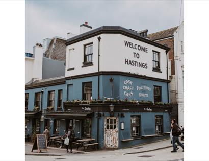 Blue-painted pub with sign reading 'Welcome to Hastings' and outdoor seating on a street corner.