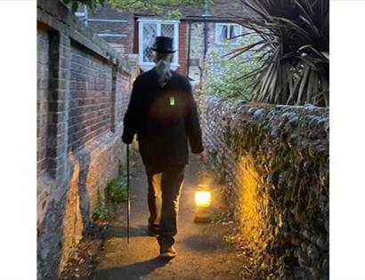 Person in dark coat and top hat walking down a narrow alley with a lantern lighting the path.