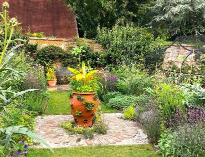 Garden scene with lush plants surrounding a large terracotta pot on a brick circle at Pashley Manor Gardens.