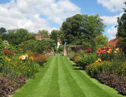 Striped lawn bordered by colourful summer flowers and greenery at Pashley Manor Gardens under a bright sky.