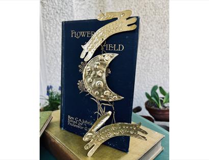 Brass ornament of two leaping animals and a crescent moon hanging in front of an old book titled ‘Flower Field’.